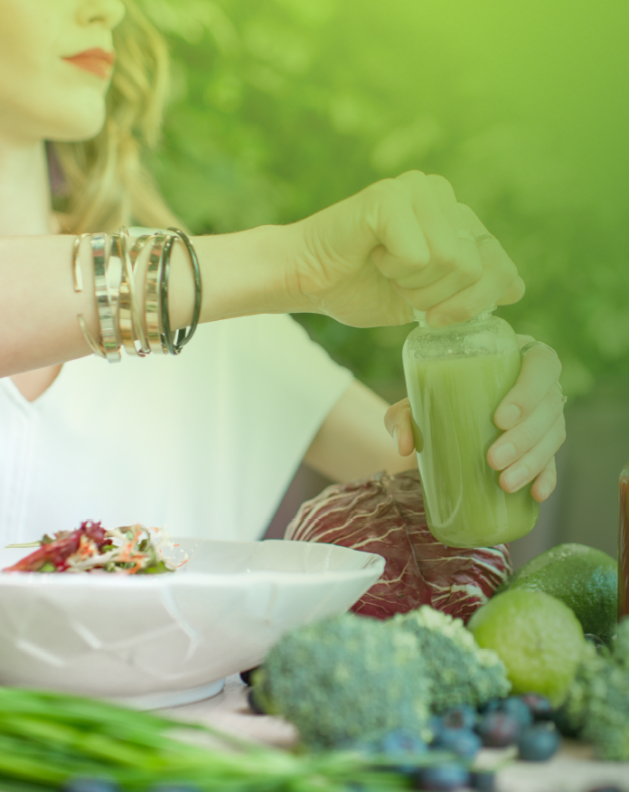 Woman opening green juice and sitting at table surrounded by fresh vegetables and a healthy salad